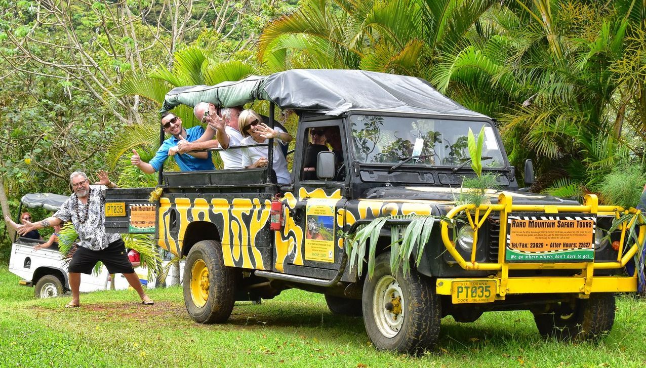 A group of people cheers from a black and yellow safari vehicle parked on grass, with one person standing outside posing.