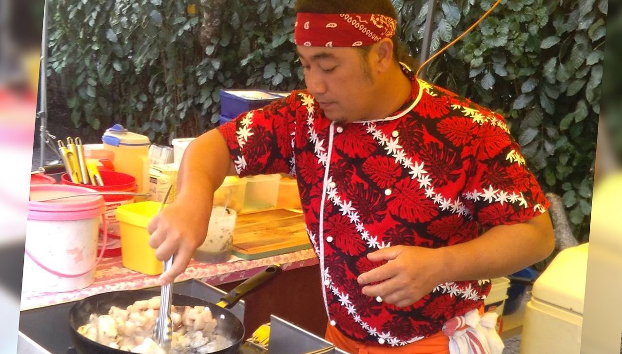 A cook in a red patterned shirt and bandana stirs food in a frying pan at a food stall.