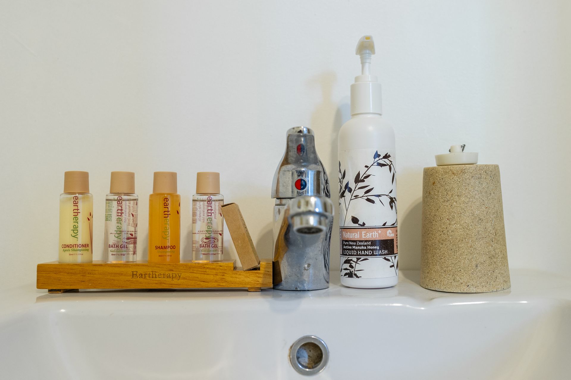 Hotel toiletries including small bottles on a wooden tray, a pump bottle, and a textured cup beside a sink faucet.