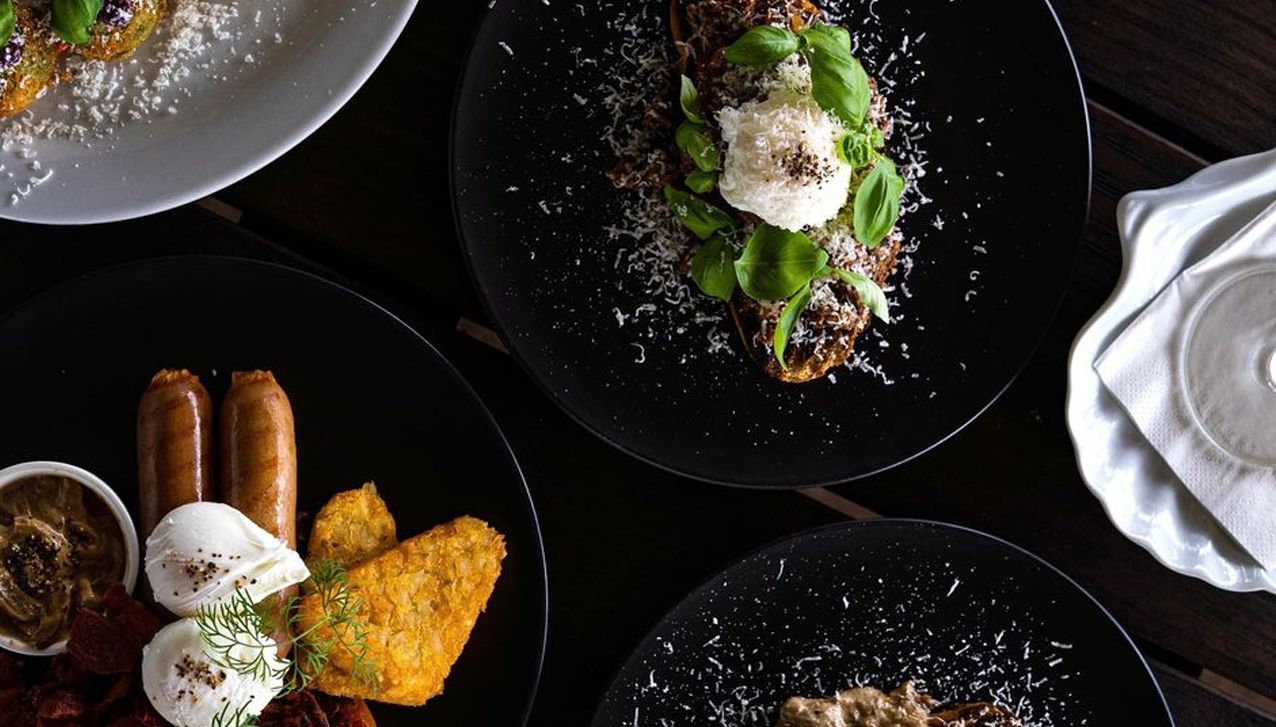 A top-down view of four black plates containing gourmet breakfast dishes, including poached eggs, sausages, and greens.