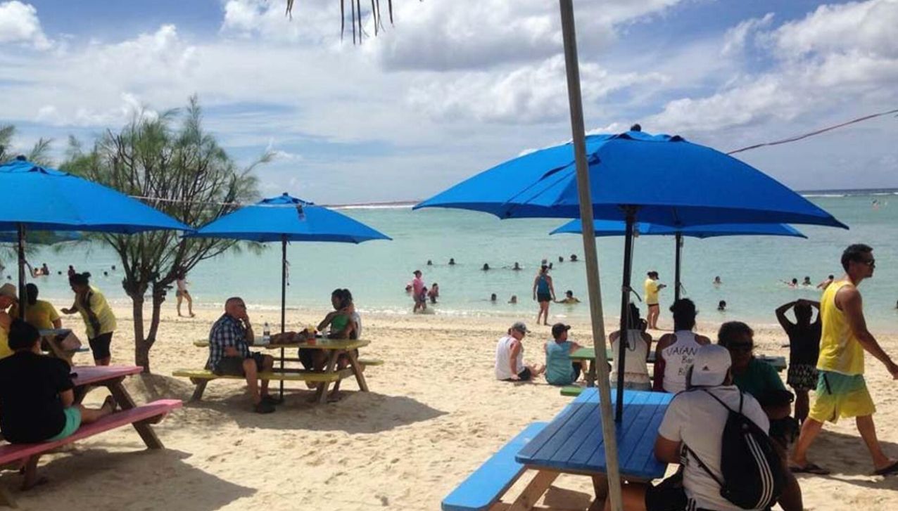 People relax at picnic tables with blue umbrellas on a sandy beach next to the ocean.