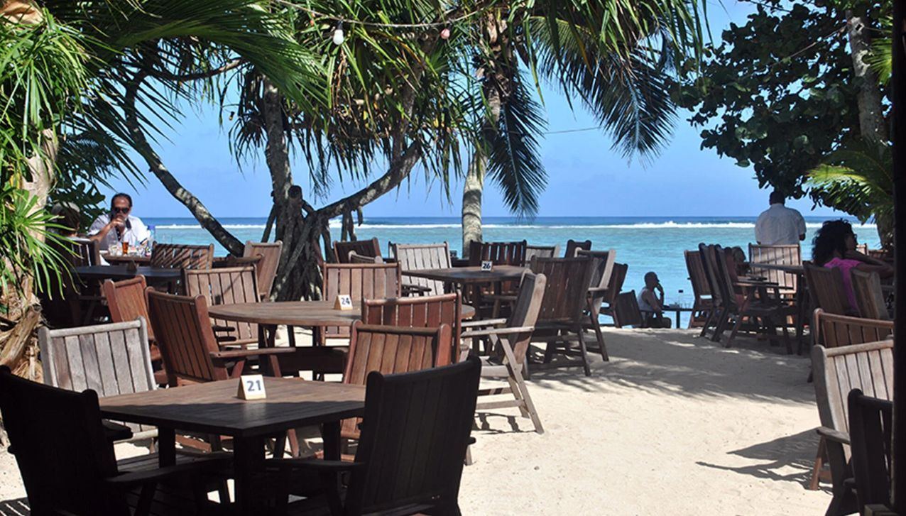 A beachfront restaurant patio with wooden tables and chairs set on sand, overlooking a bright blue ocean under palm trees.