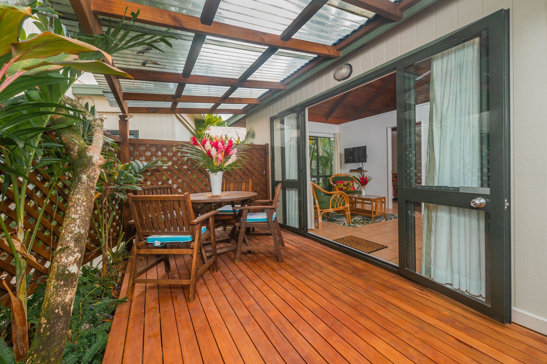 A wooden deck with a dining set under a covered pergola, viewed through an open glass sliding door from an indoor room.