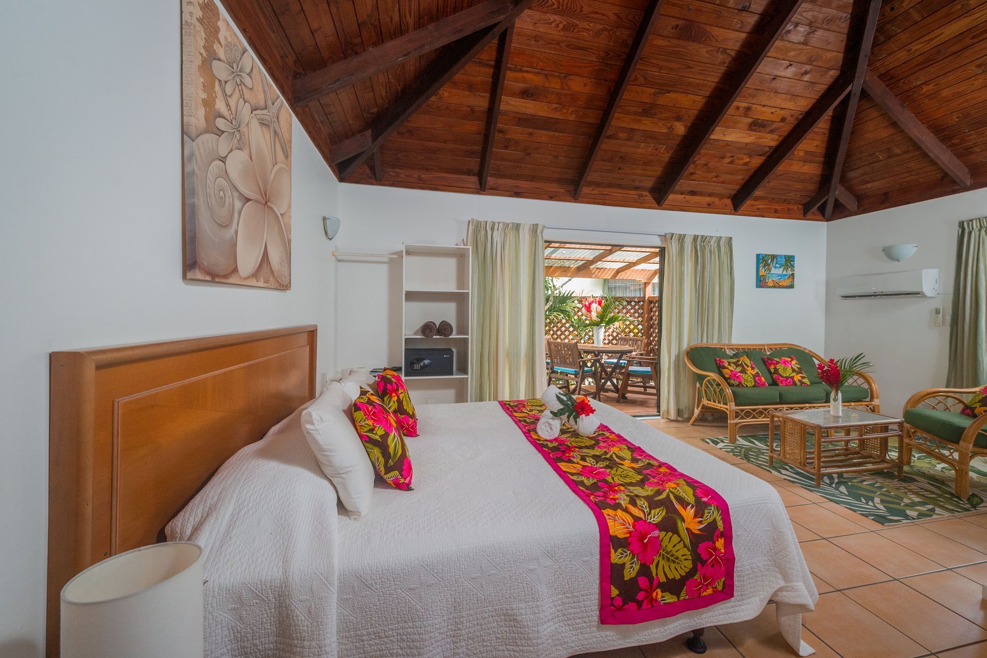 A bright hotel room with a wooden vaulted ceiling, a king bed featuring a floral runner, and a seating area by a terrace.