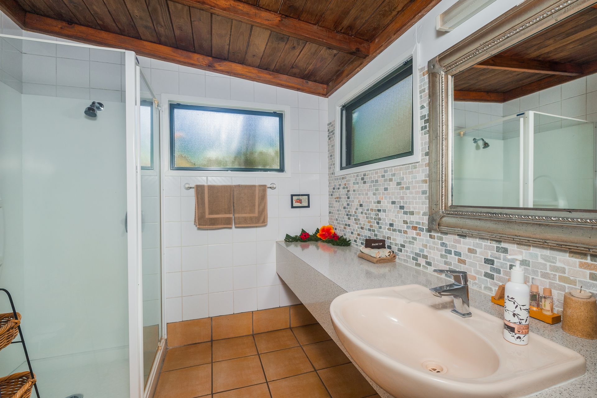 A tiled bathroom features a glass-enclosed shower, a long countertop with a sink, patterned walls, and wood ceiling beams.