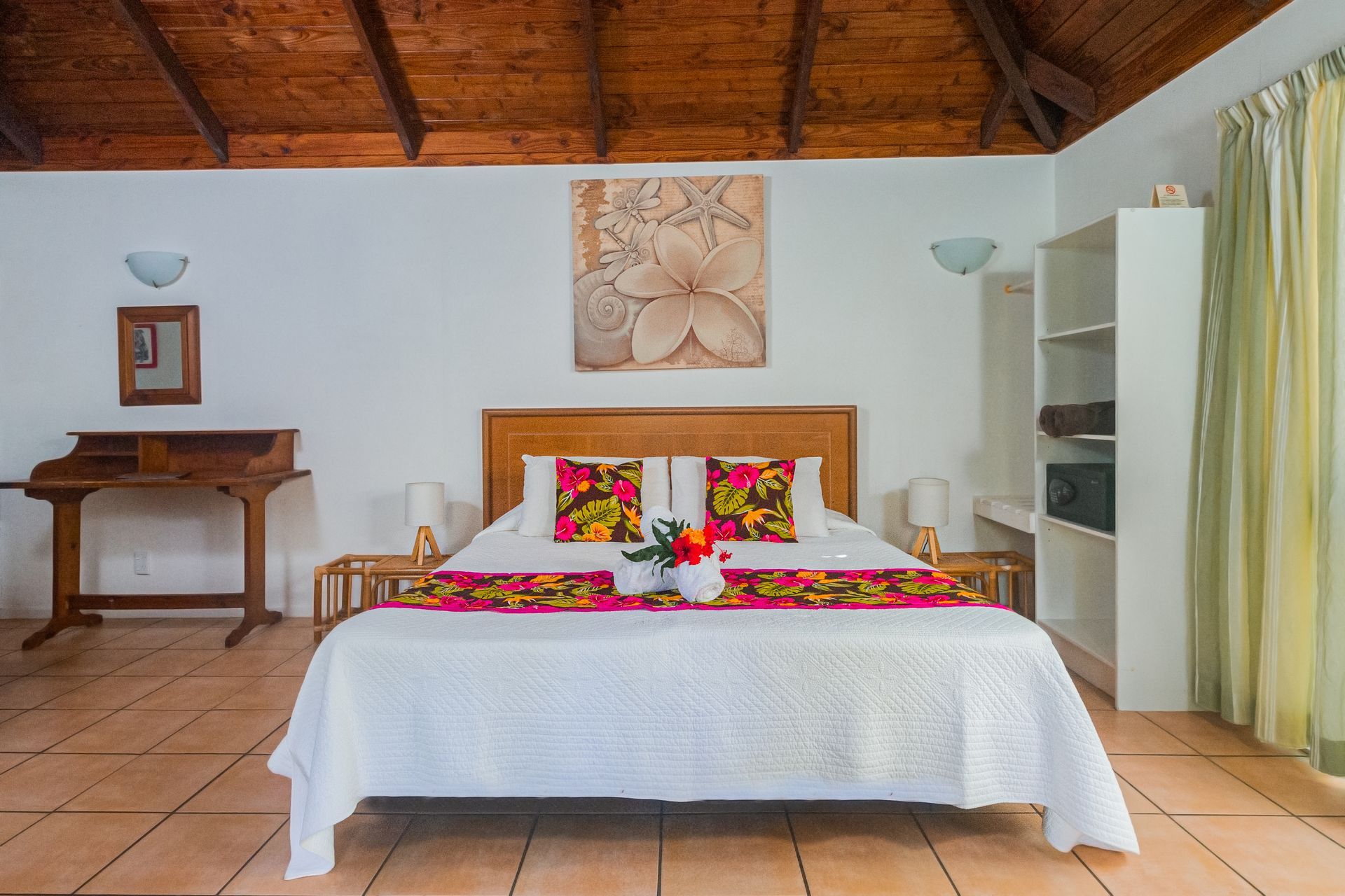 A bedroom with a king-sized bed, white linens, floral accent pillows, wood ceiling, and a desk against a white wall.
