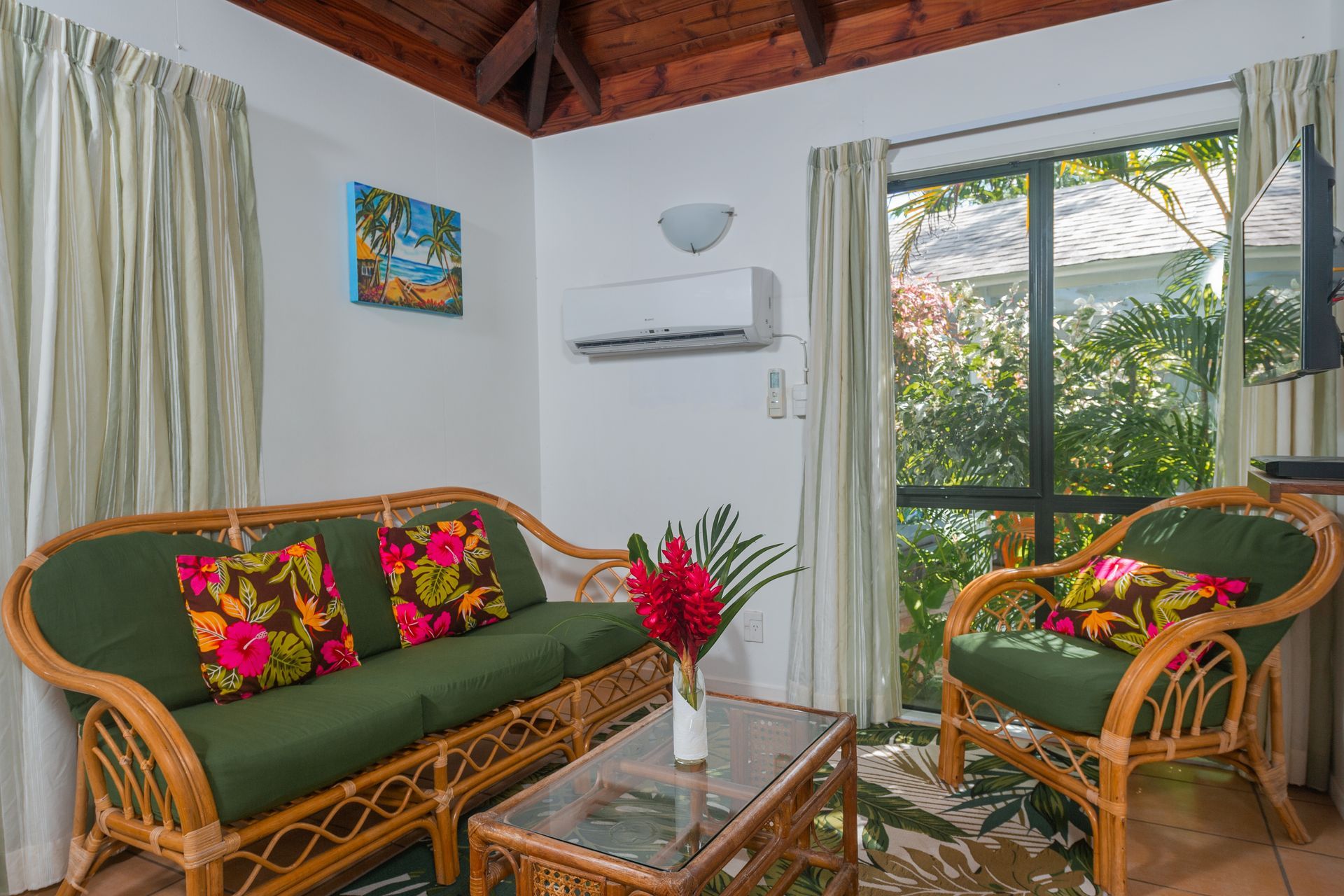 A cozy seating area with wicker furniture, green cushions with floral pillows, a glass coffee table, and large windows.