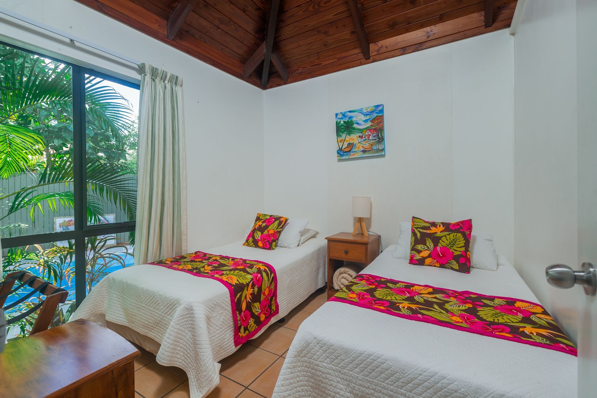 A bright bedroom with two twin beds, colorful floral runners, a wooden nightstand, and a window overlooking greenery.