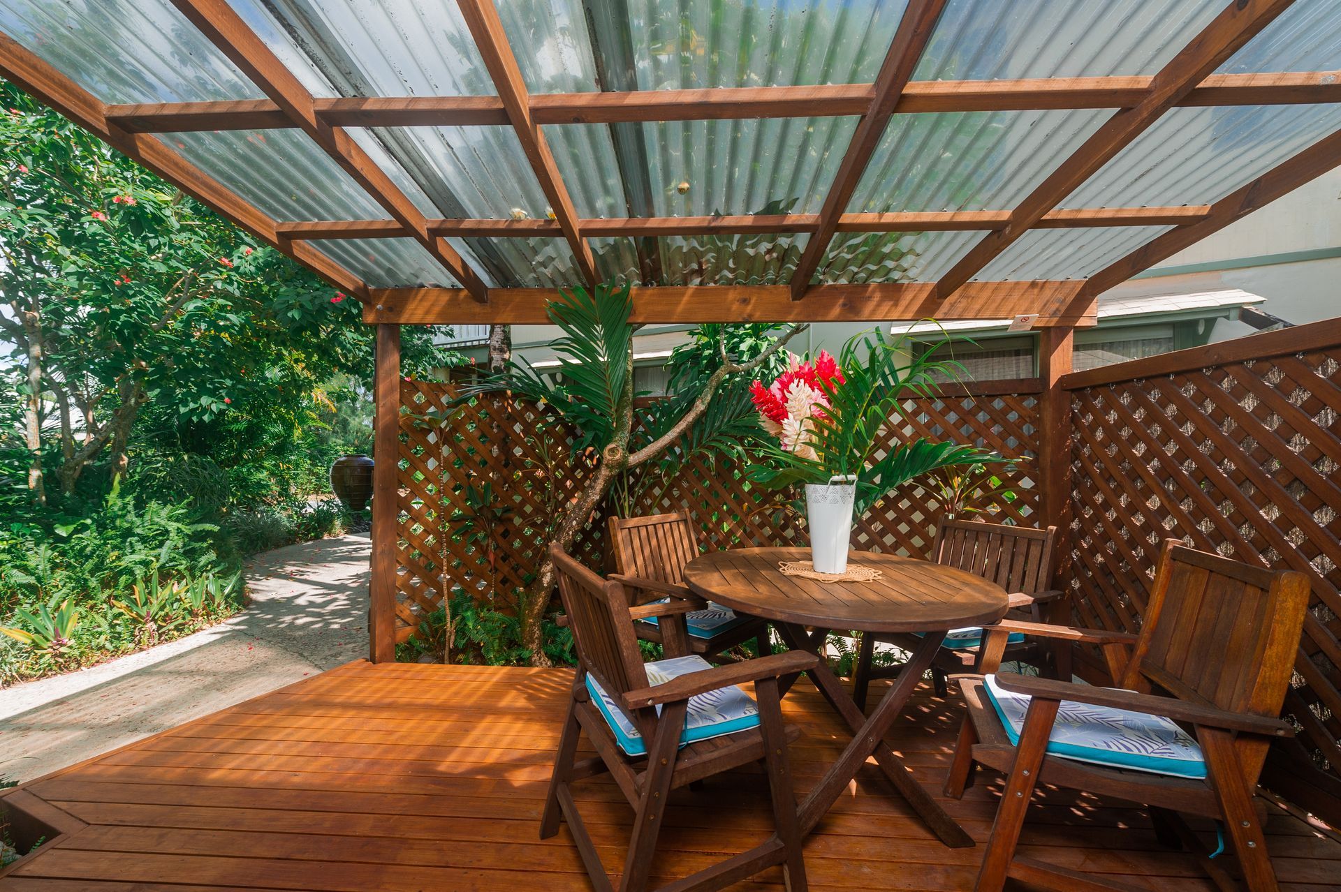 Wooden patio set with chairs and a table under a corrugated roof on a deck, surrounded by lattice fencing and greenery.