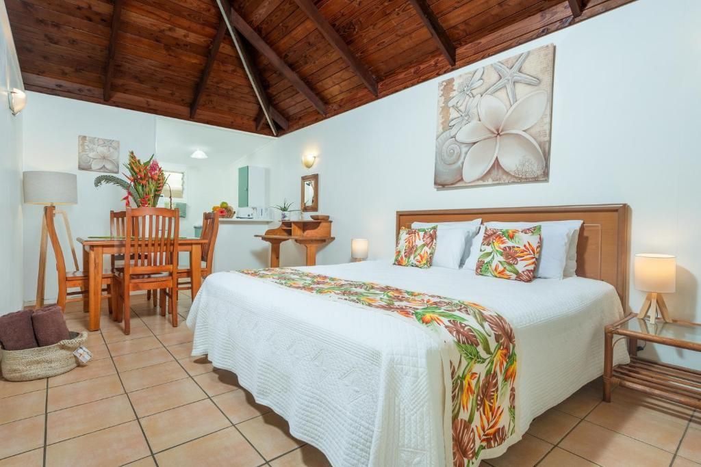 A tropical hotel room with a large bed, wooden vaulted ceiling, dining area, and floral decor.