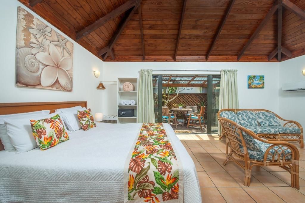 A hotel room with a white bed, floral accents, wooden vaulted ceiling, and a sitting area opening to a terrace.