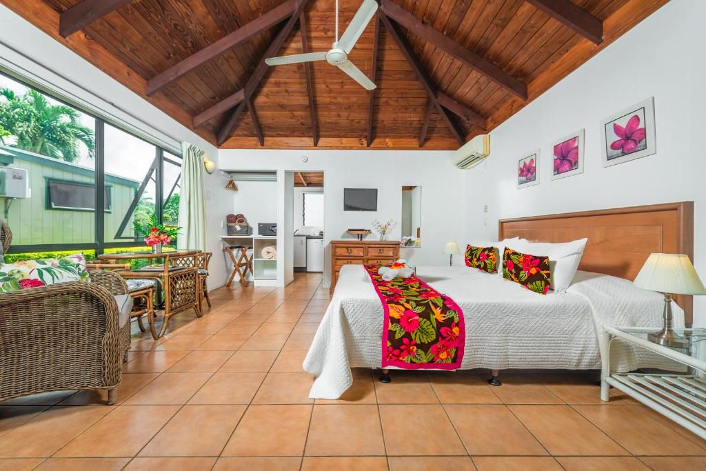 A bright, airy hotel room featuring a large bed with a floral runner, wicker furniture, and a high, wooden beamed ceiling.