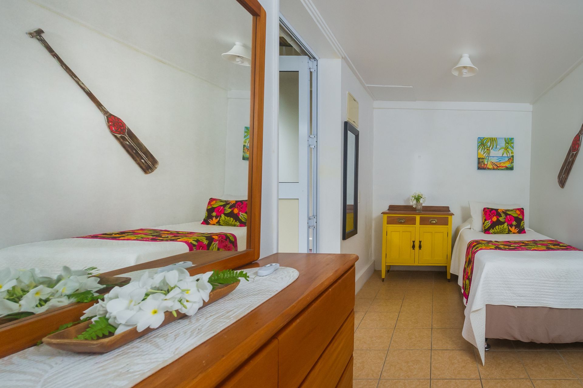 A bedroom with twin beds, a yellow cabinet, and a wooden dresser with white flowers, viewed through a mirror.