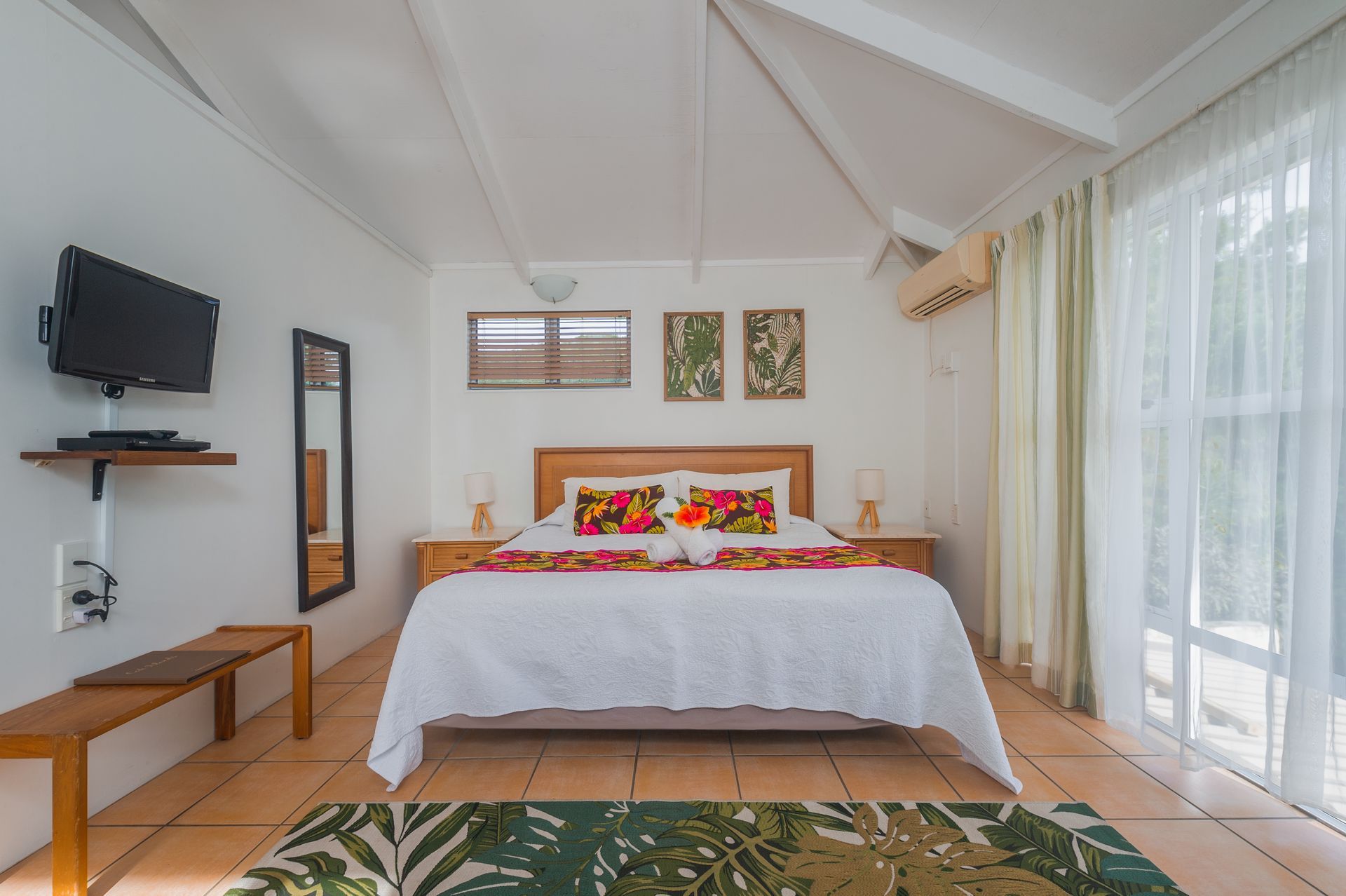 A bright bedroom with a queen bed featuring a white quilt and floral pillows, tiled floors, and large glass doors.