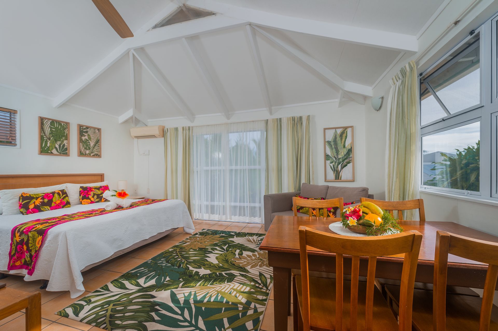 A tropical-themed hotel room with a bed, dining set, and vaulted ceiling, decorated in leafy green and floral patterns.