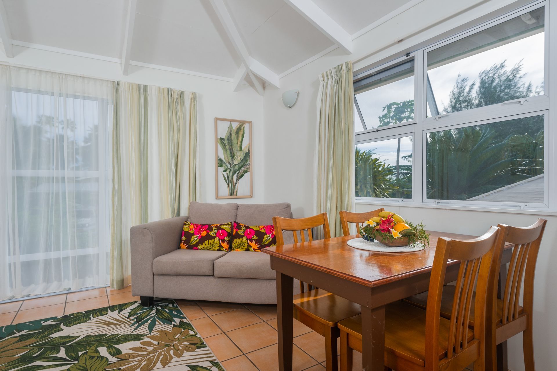 A sunlit room with a light gray sofa, a wooden dining set, a patterned rug, and curtains over sliding glass doors.