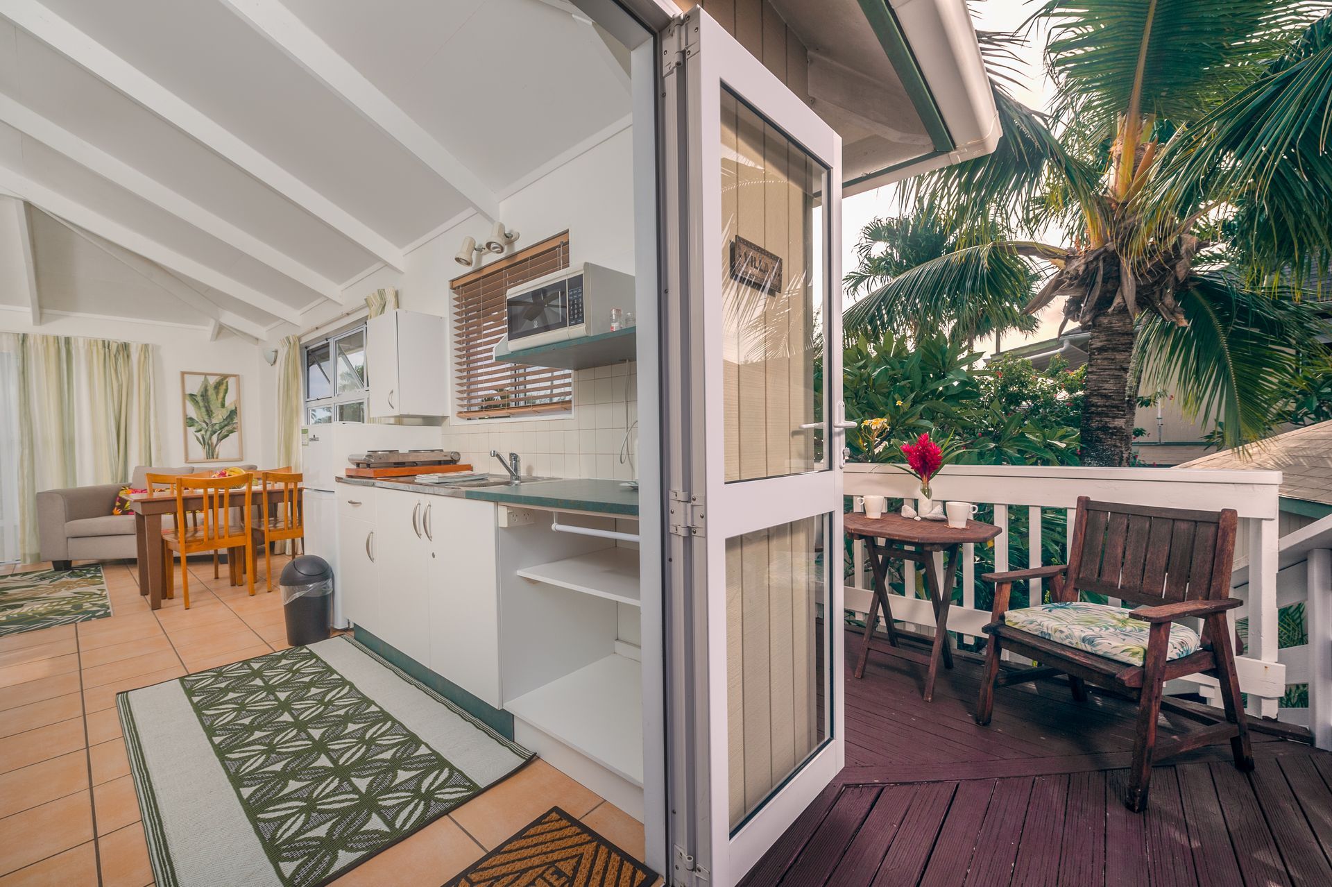 A view from an indoor kitchen through an open door onto a wooden deck with chairs, overlooking lush tropical trees.