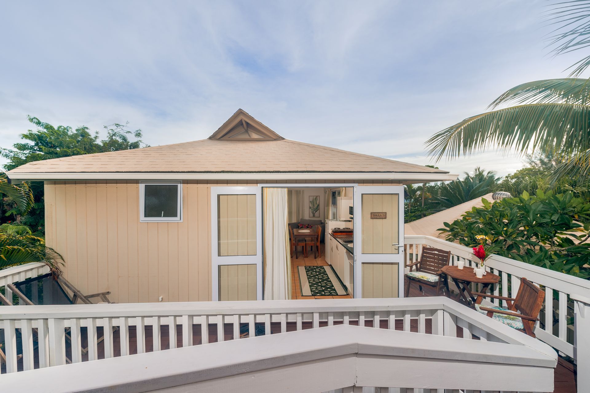 A light beige tropical cottage with an open doorway leading to a dining area, surrounded by a white deck and lush greenery.