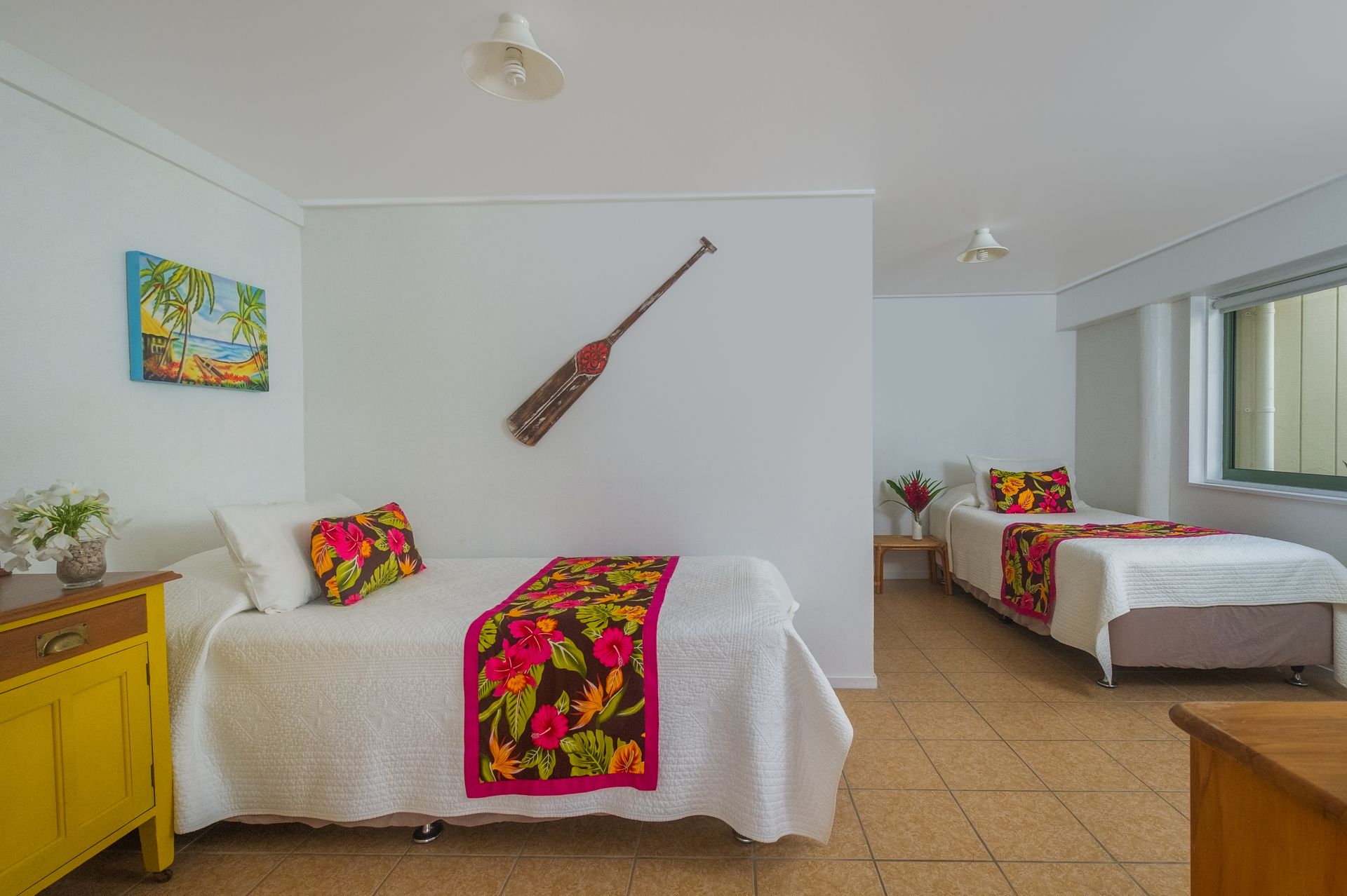 Two twin beds with tropical-print runners in a brightly lit bedroom with white walls and a decorative wooden oar.