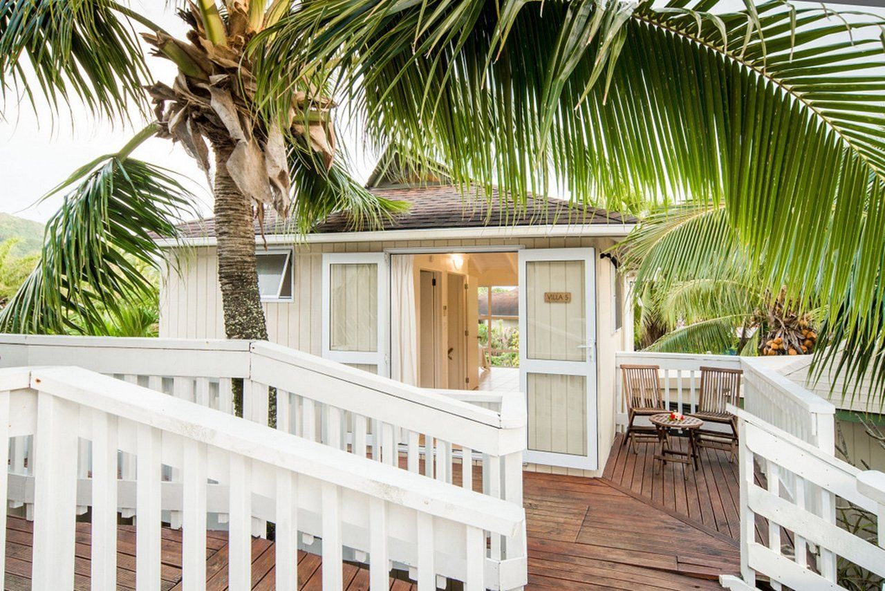 White wooden cottage with a ramp entrance, palm trees, and a deck with chairs in a lush, tropical setting.