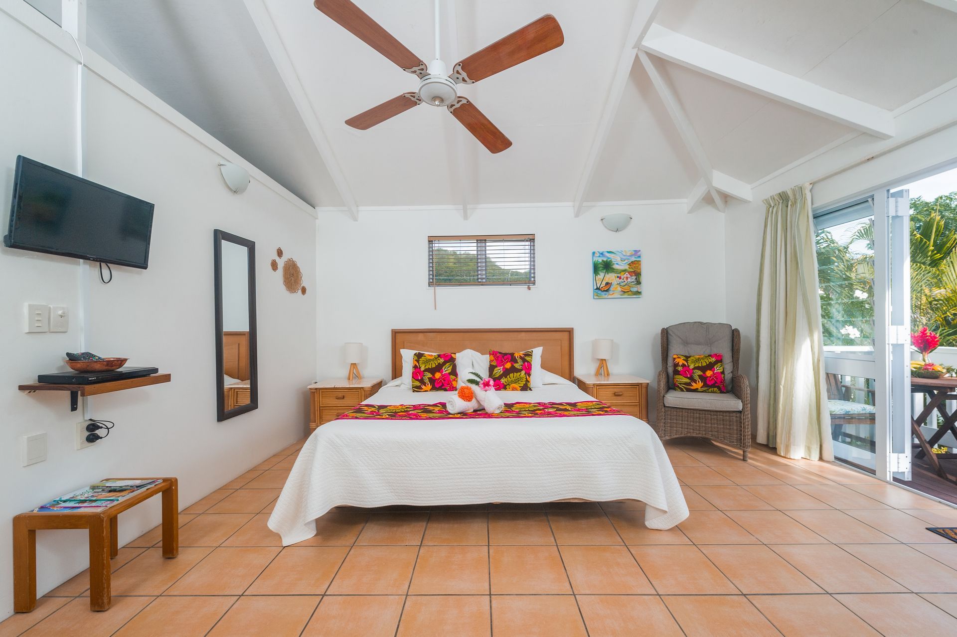 A bright bedroom with a queen bed, tiled floors, a ceiling fan, a wall-mounted TV, and a glass door leading to a patio.
