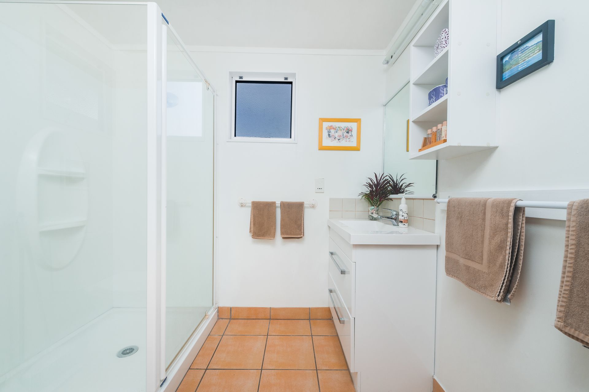 A bright bathroom with a glass-enclosed shower, a white vanity, tan floor tiles, and beige towels hanging on the walls.
