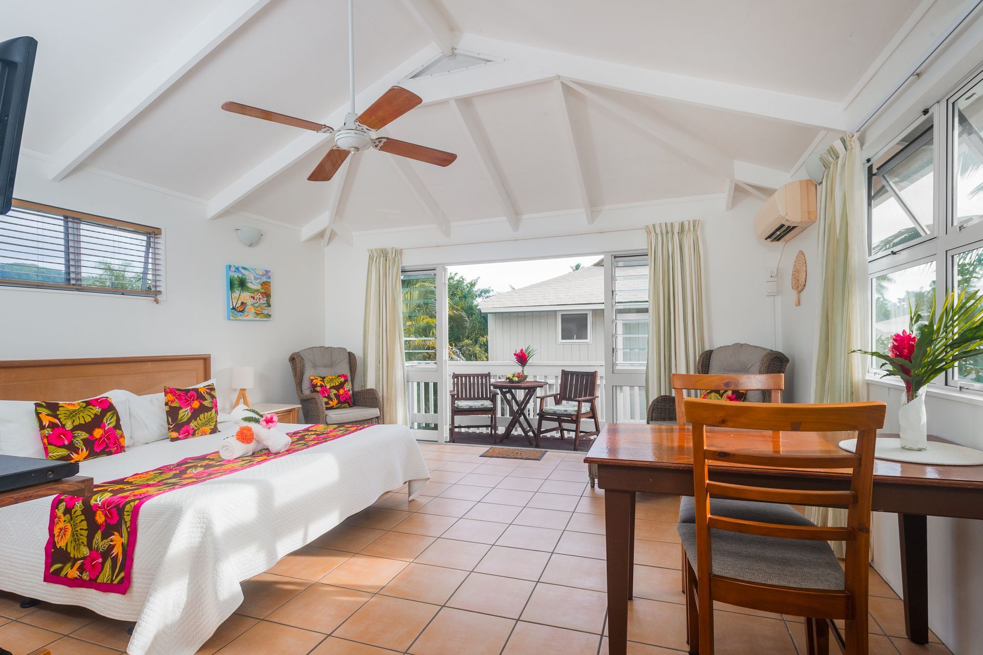 A bright, airy hotel room with white walls, tiled floors, a king bed with floral runners, and a dining area with a balcony.