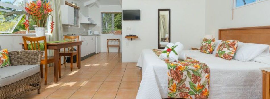 A hotel room with a queen bed, a dining table and chairs, a small kitchenette, and wicker furniture on a tiled floor.