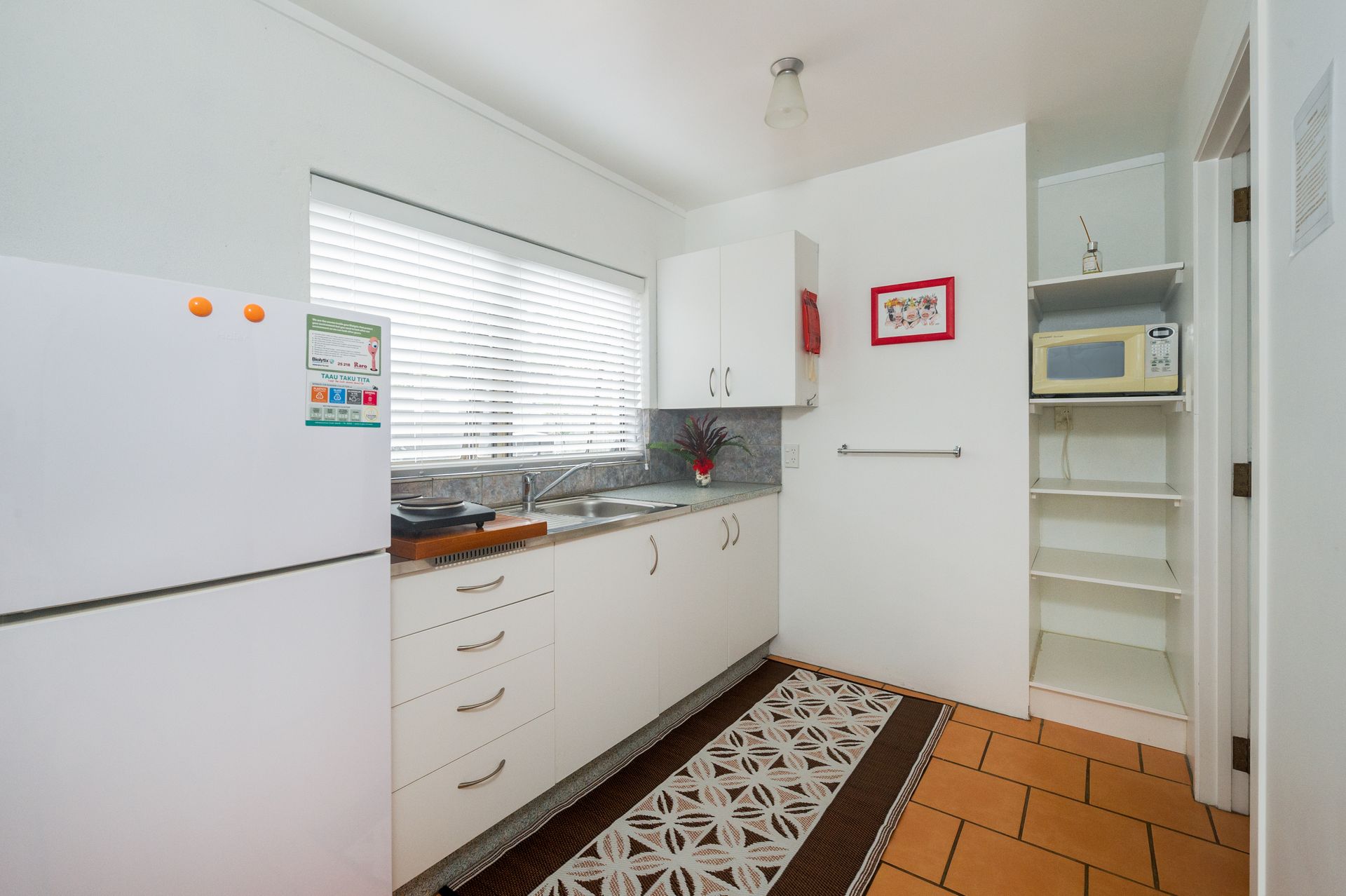 A white kitchen featuring cabinets, a refrigerator, a window with blinds, a microwave, and a patterned rug on tiled floors.
