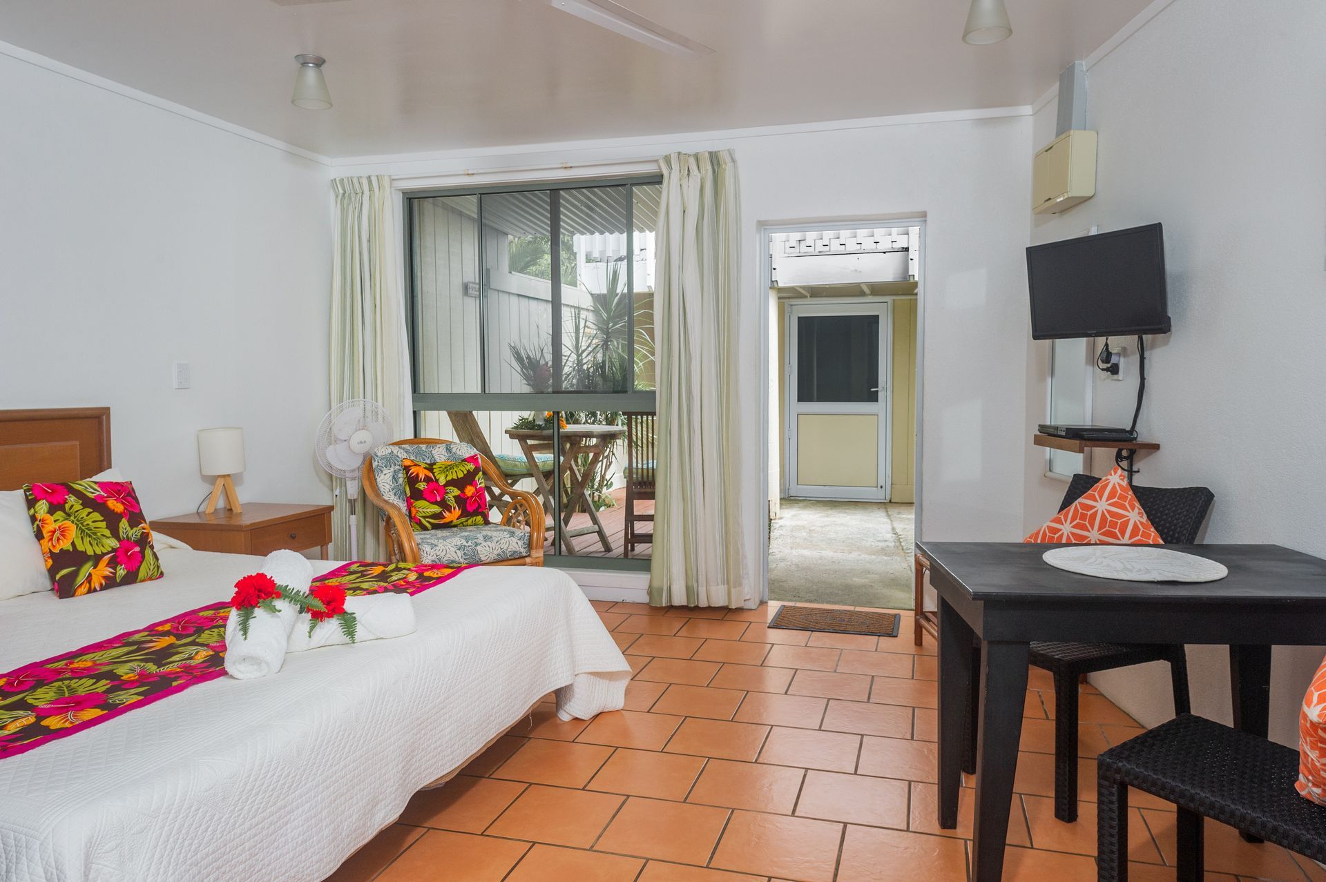 A bright hotel room with a bed featuring a colorful runner, a small table, a wall-mounted TV, and a patio door view.