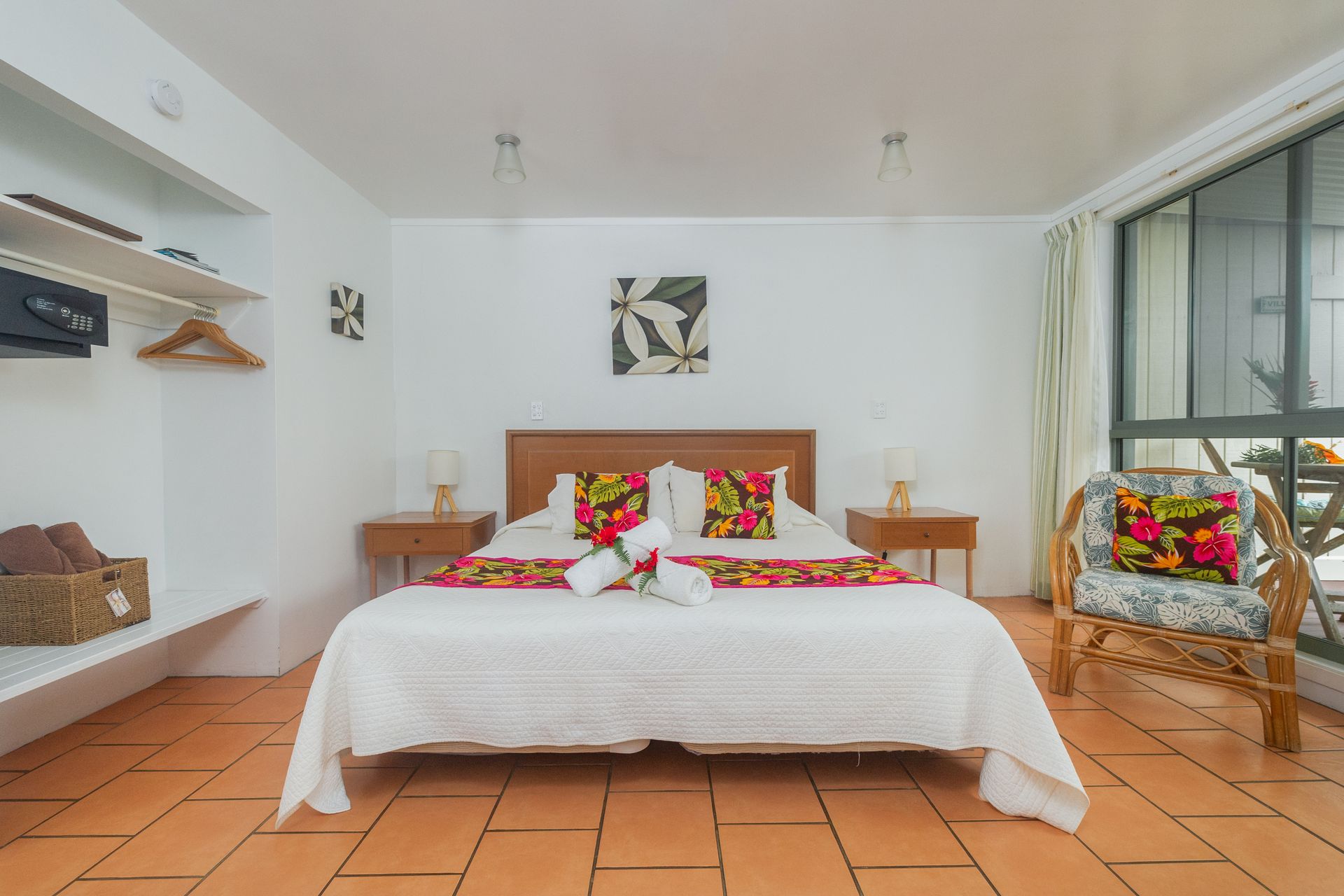 A bright bedroom with a queen bed featuring a white quilt, floral accents, wood nightstands, and a rattan chair.