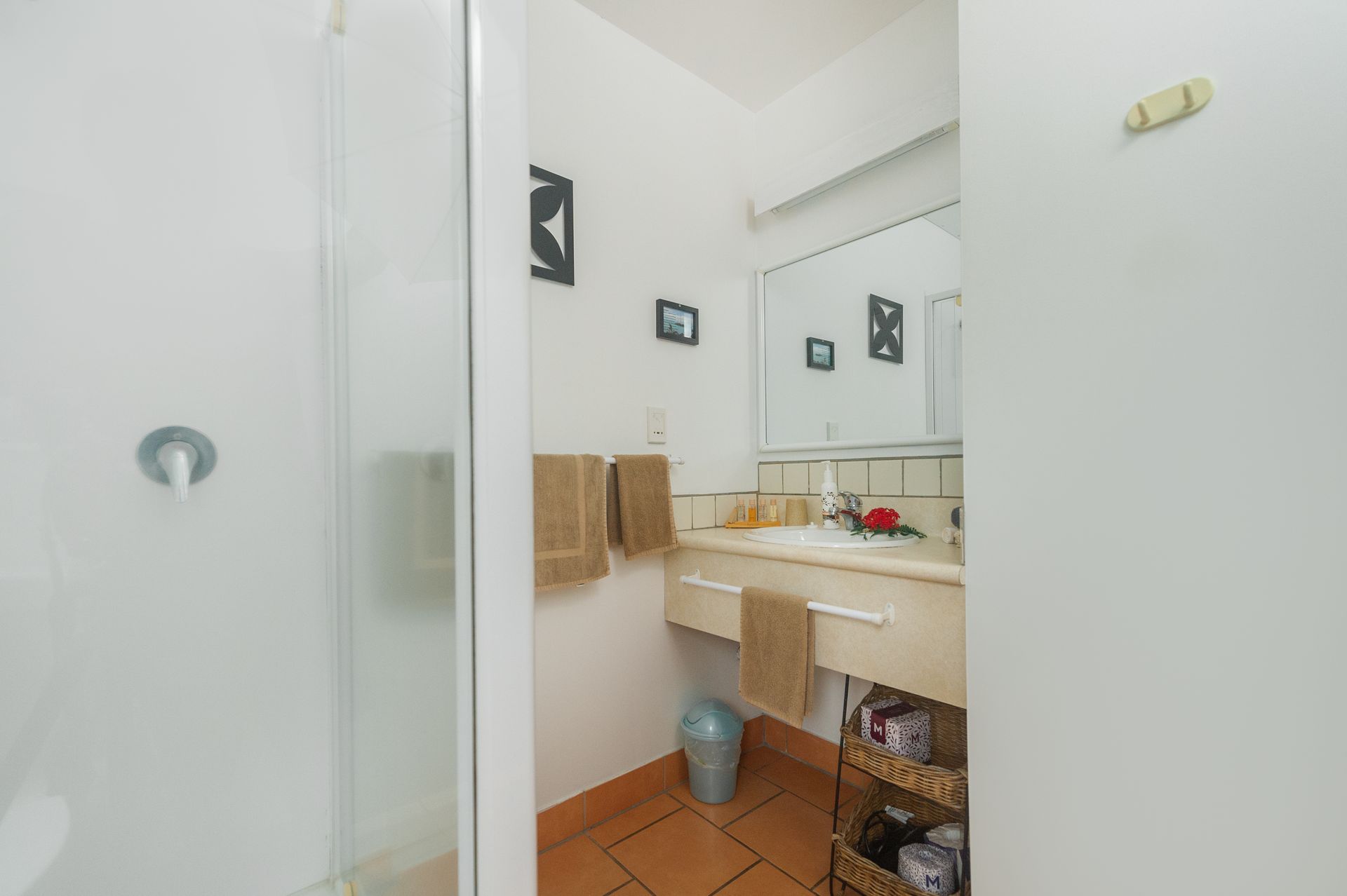 A small bathroom with a glass-enclosed shower, a vanity sink, mirror, neutral towels, and terracotta floor tiles.