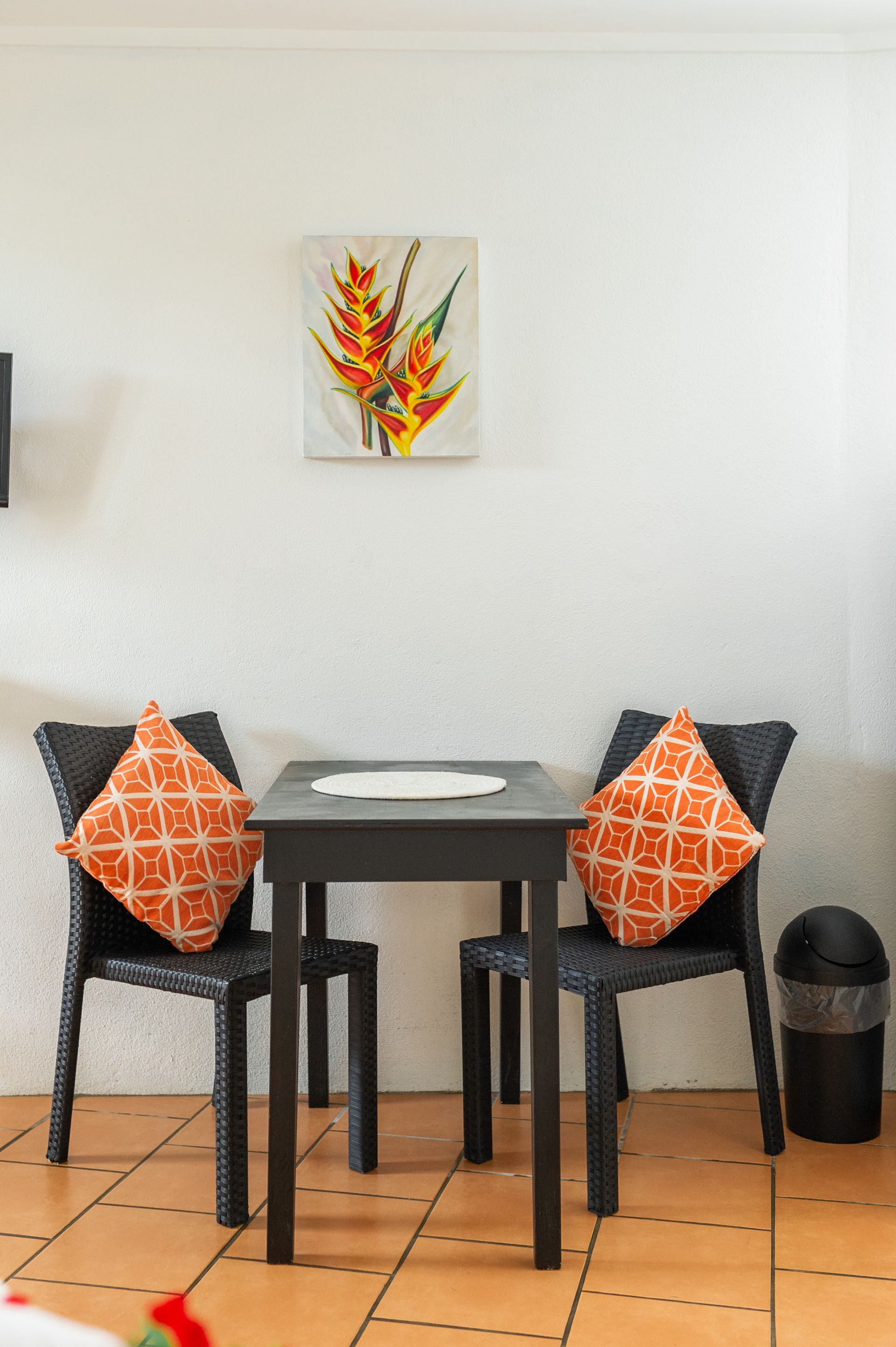 A small black table with two woven chairs featuring orange patterned cushions, set against a white wall with floral art.