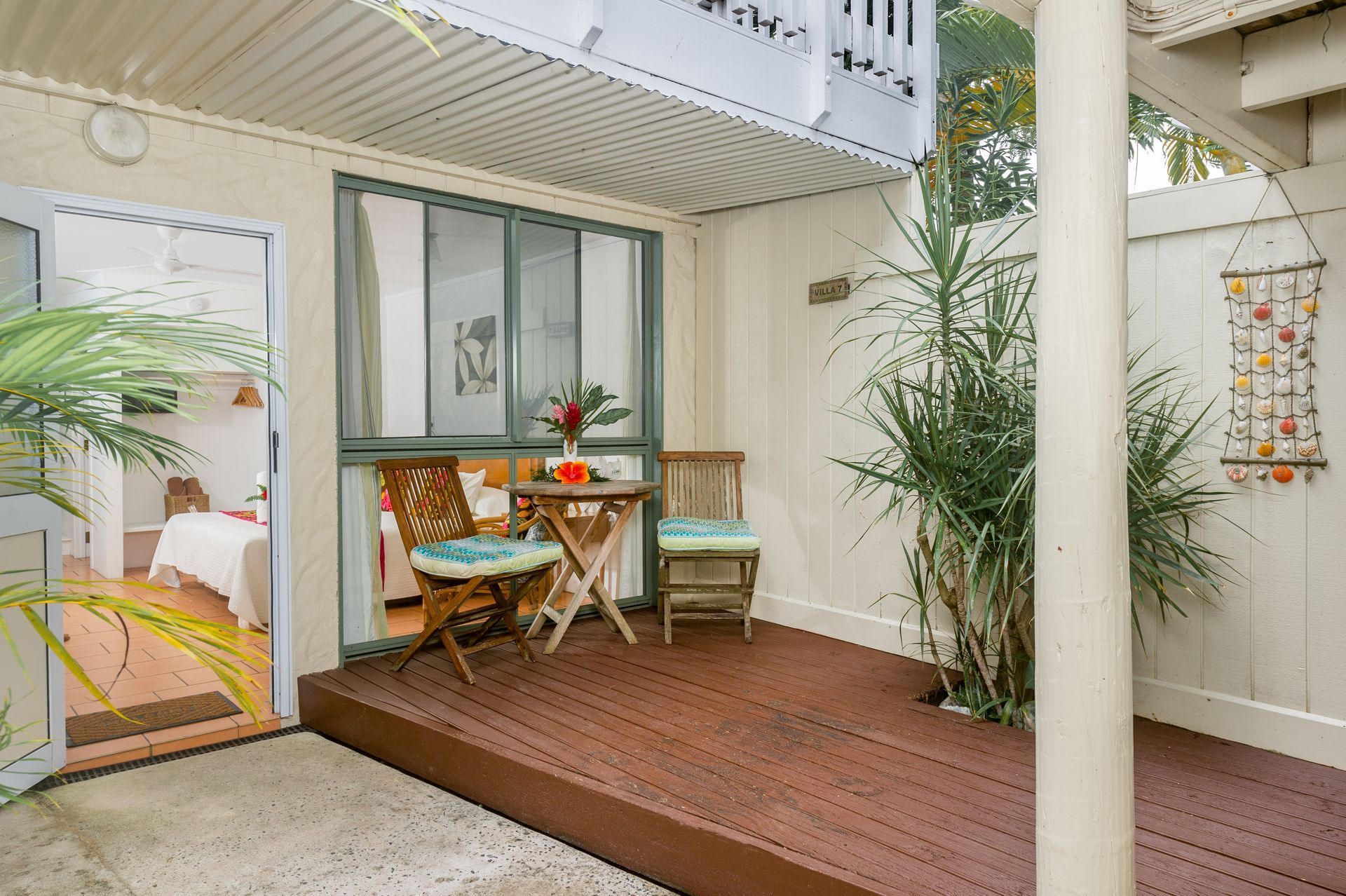 A wooden deck patio features two chairs and a small table, with a glass door leading to a bedroom and potted greenery.