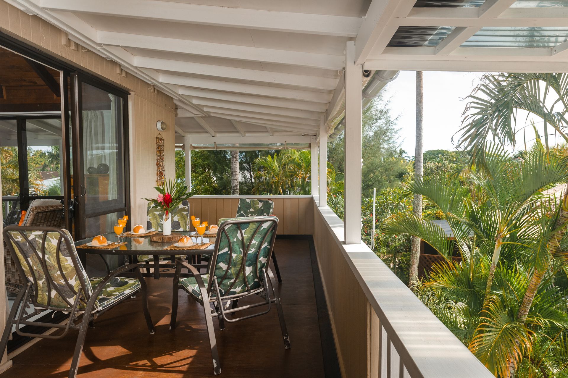 An outdoor dining set with four patterned chairs sits on a covered, wooden porch overlooking lush tropical greenery.
