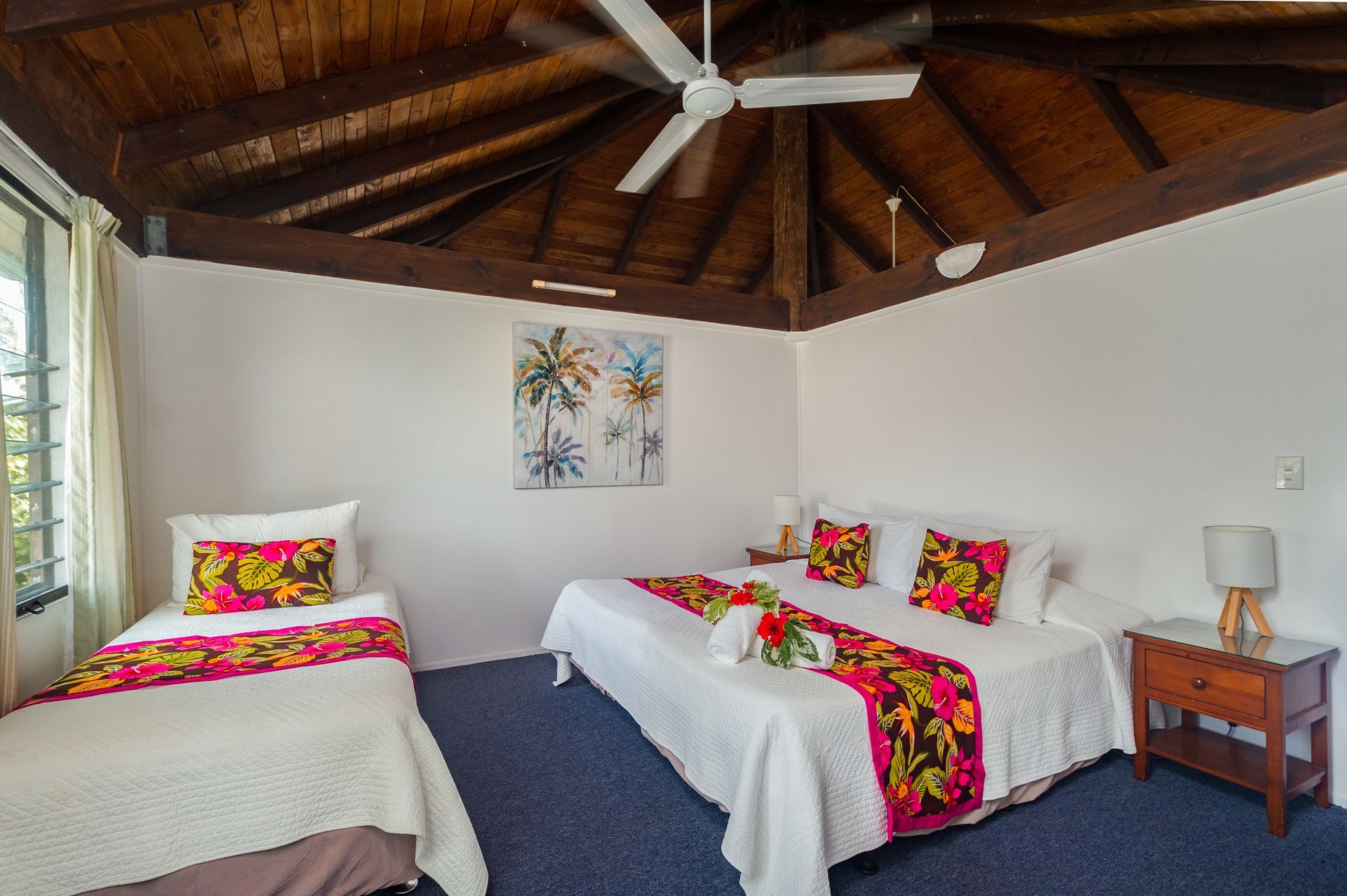 A bedroom with a king bed and a twin bed, white linens with floral runners, wooden vaulted ceiling, and blue carpet.