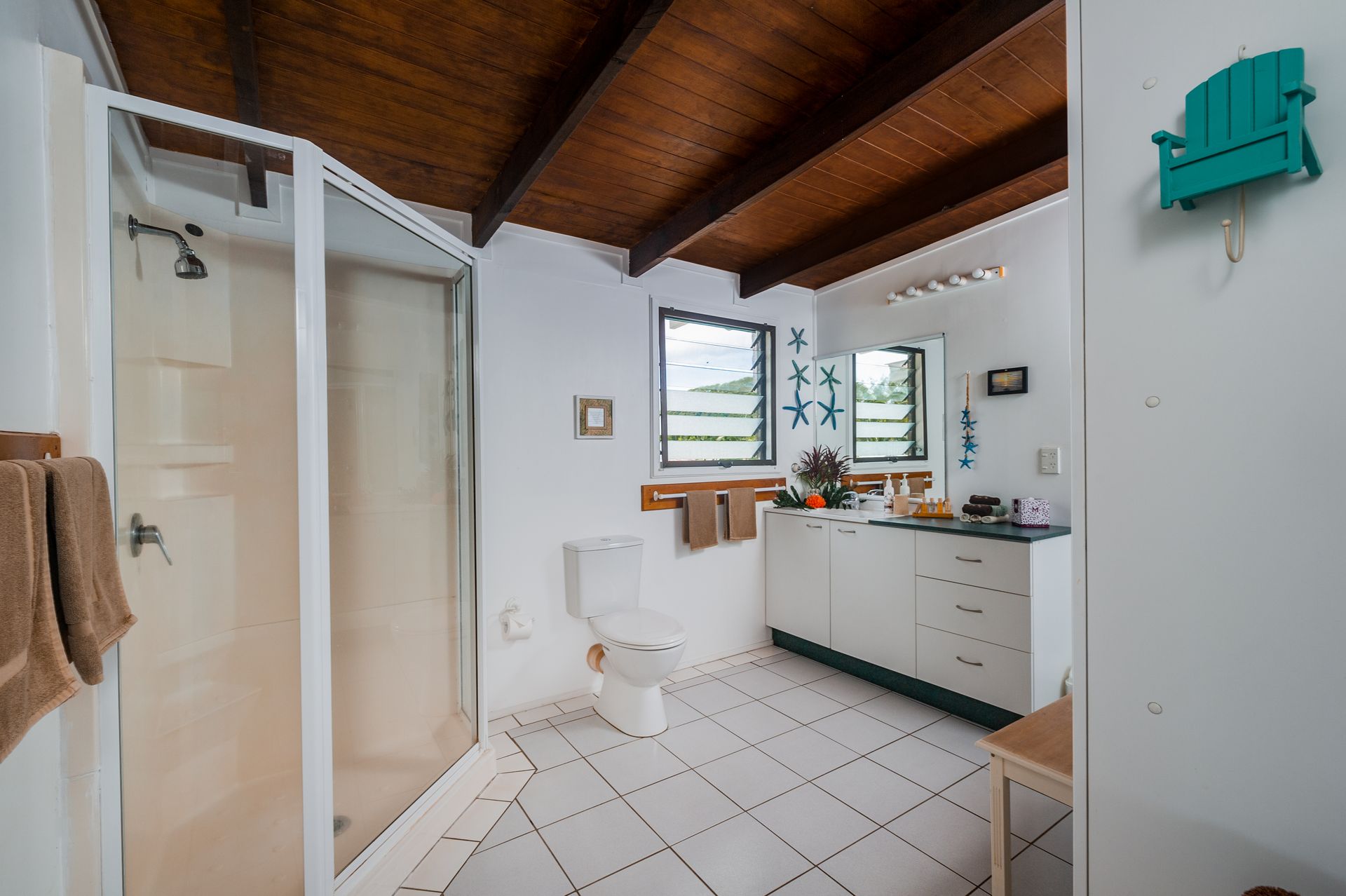 A bathroom with white tiled floors, a corner shower, a toilet, a white vanity, and a wooden vaulted ceiling.