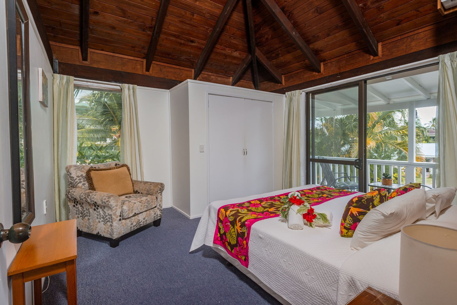A hotel bedroom with a bed featuring a floral runner, a patterned armchair, a wood-beamed ceiling, and a glass door.