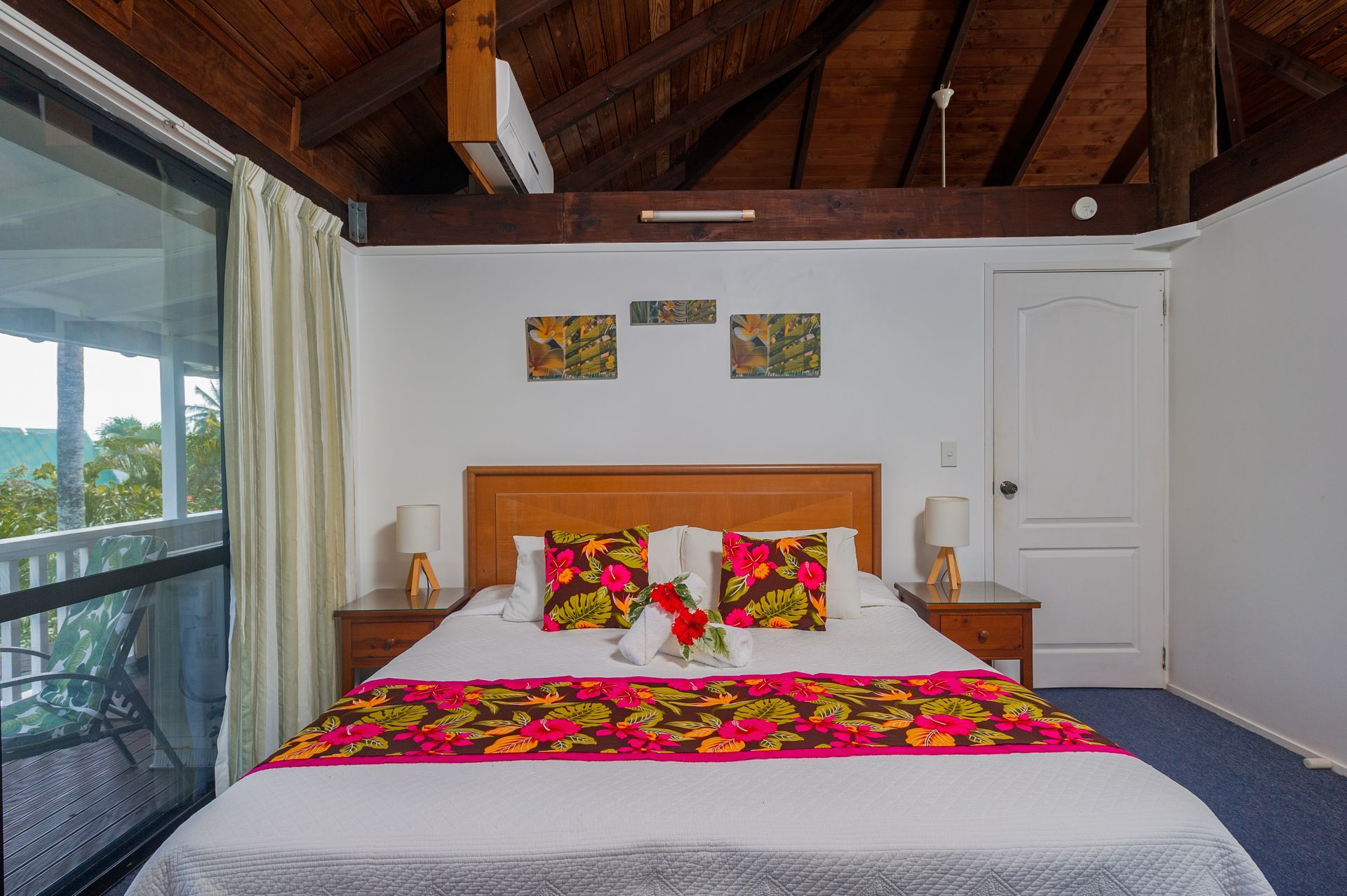 A bedroom with a queen bed featuring floral bedding, wooden ceiling beams, white walls, and a sliding glass door.