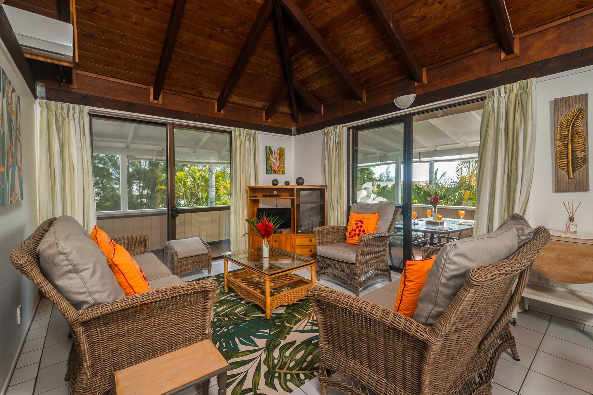Sunlit living room with vaulted wood ceilings, wicker furniture, orange accent pillows, and large glass doors.