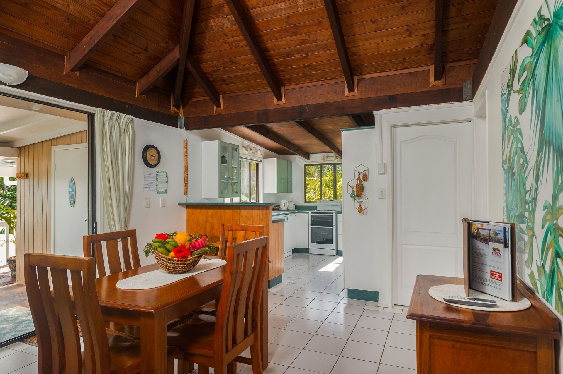 A dining area with a wooden table and chairs under a vaulted wood-beam ceiling, next to an open kitchen with white tiles.