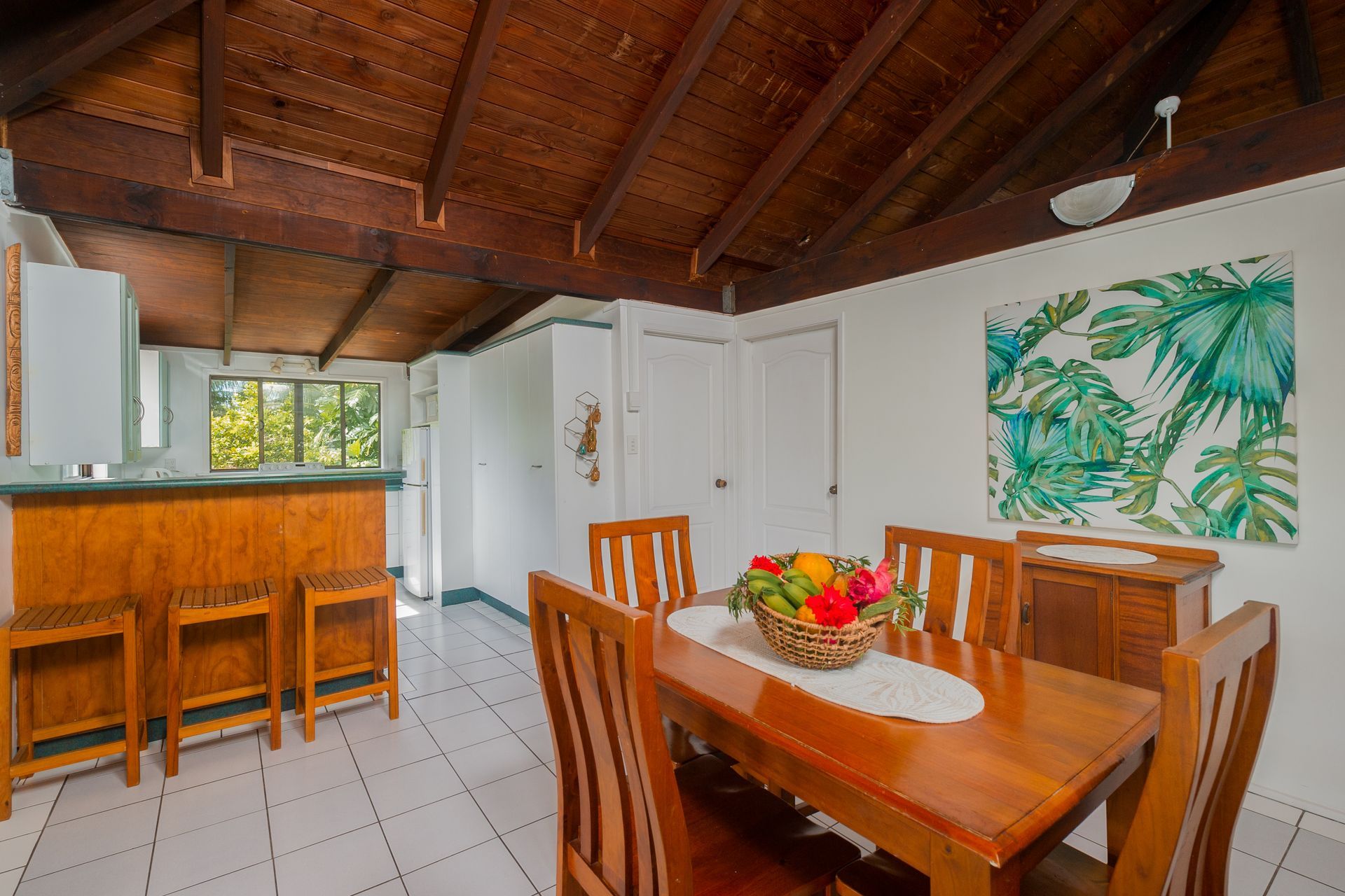 A dining room with a wooden table, chairs, fruit bowl, and high-top kitchen counter under a vaulted wooden ceiling.