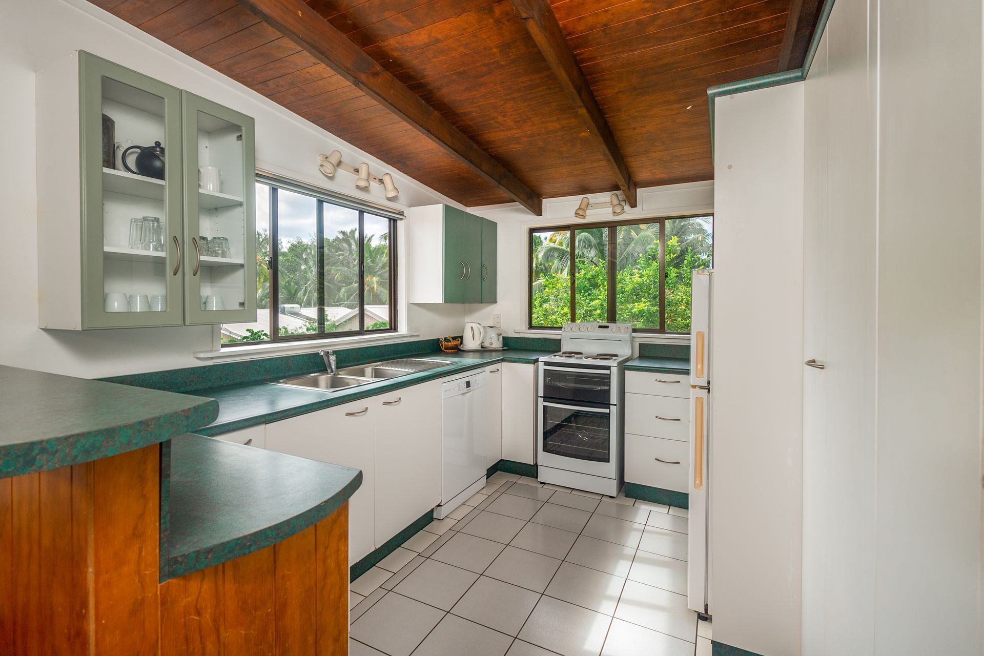 Kitchen with white cabinets, green countertops, wooden vaulted ceiling, tiled floor, and two windows.