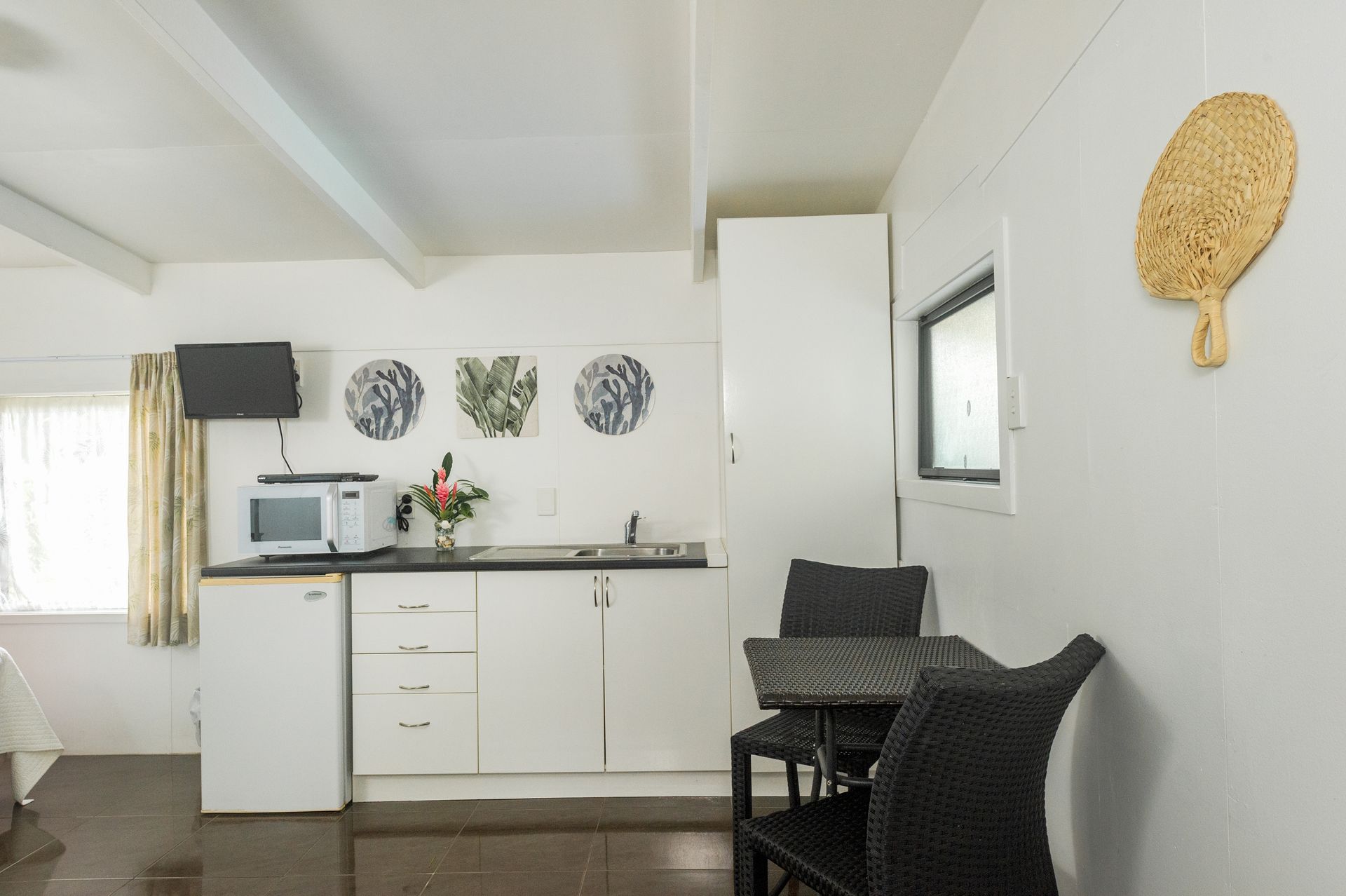 A small kitchenette with white cabinets, a microwave, mini-fridge, and a small table with two black chairs.