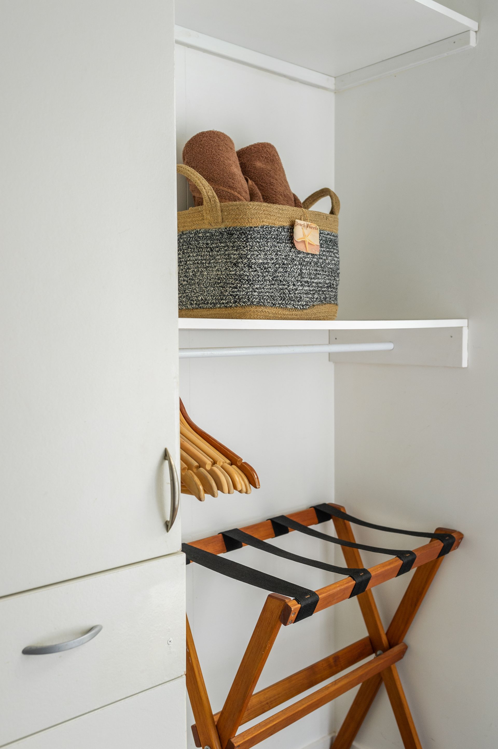 A white closet with a woven basket on a shelf, wooden hangers, and a wooden luggage rack on the floor.