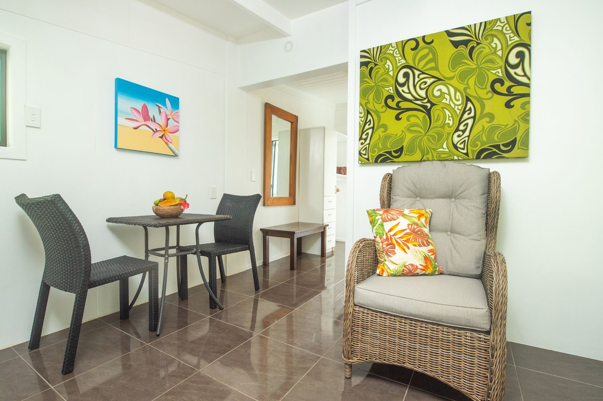 A room with a small bistro table and two black chairs, next to an armchair with a patterned pillow and green wall art.