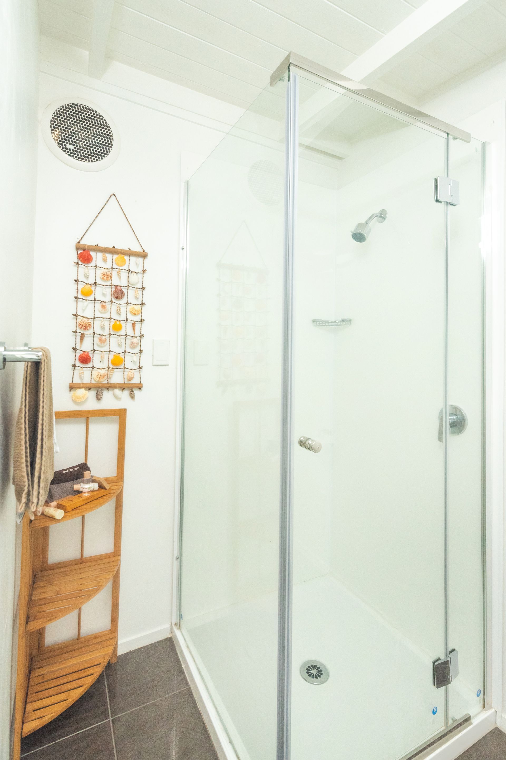 A corner shower stall with glass doors, next to a three-tiered wooden corner shelf and a hanging wall decoration.