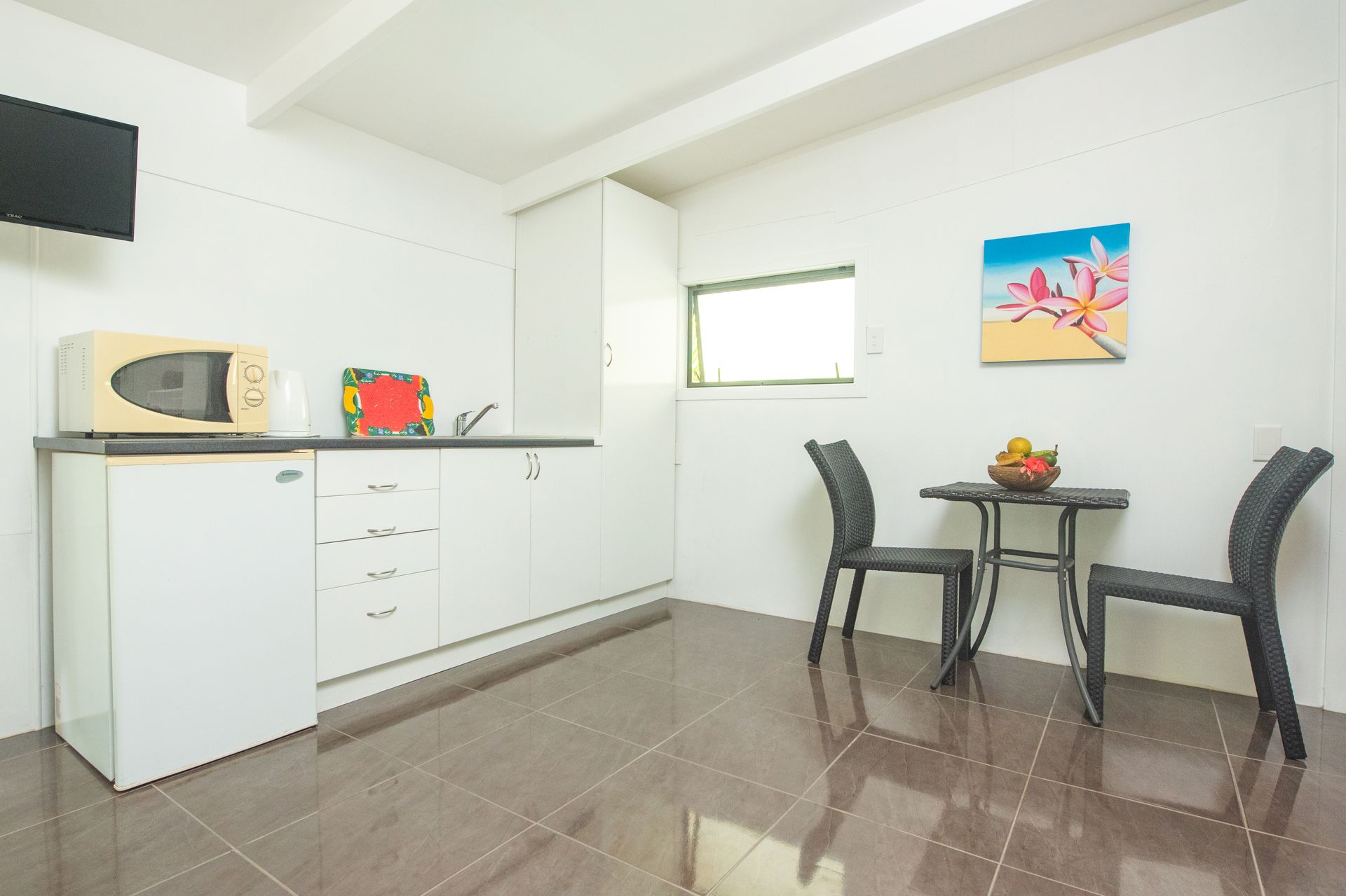 A small kitchenette with a microwave, white cabinets, tiled floors, and a small dining table with two chairs.