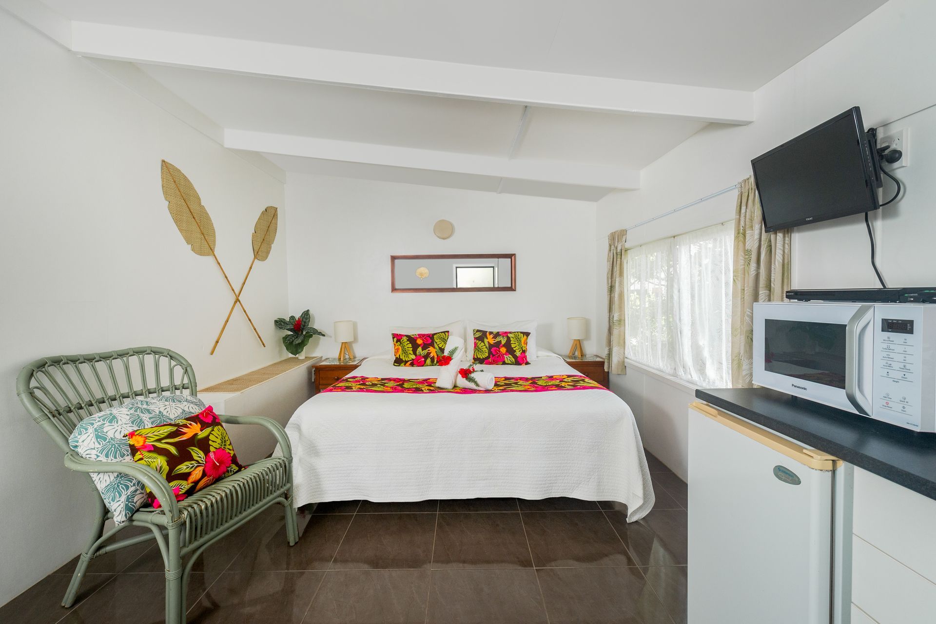 A tidy hotel room with a made bed, a green wicker chair, a microwave, and a small refrigerator under a wall-mounted TV.