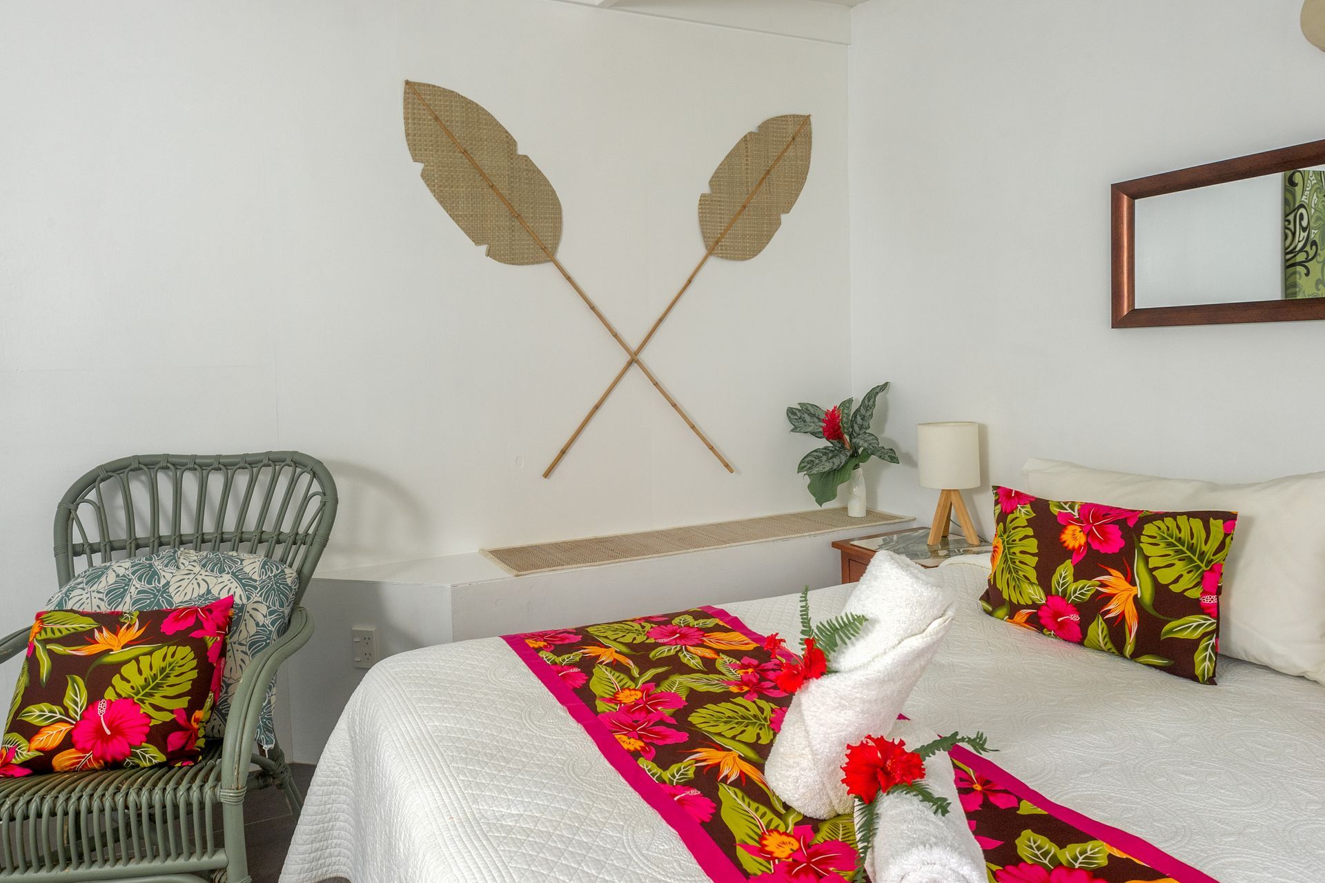 A hotel room featuring a bed with a colorful floral runner, decorative pillows, and crossed leaf wall art above it.